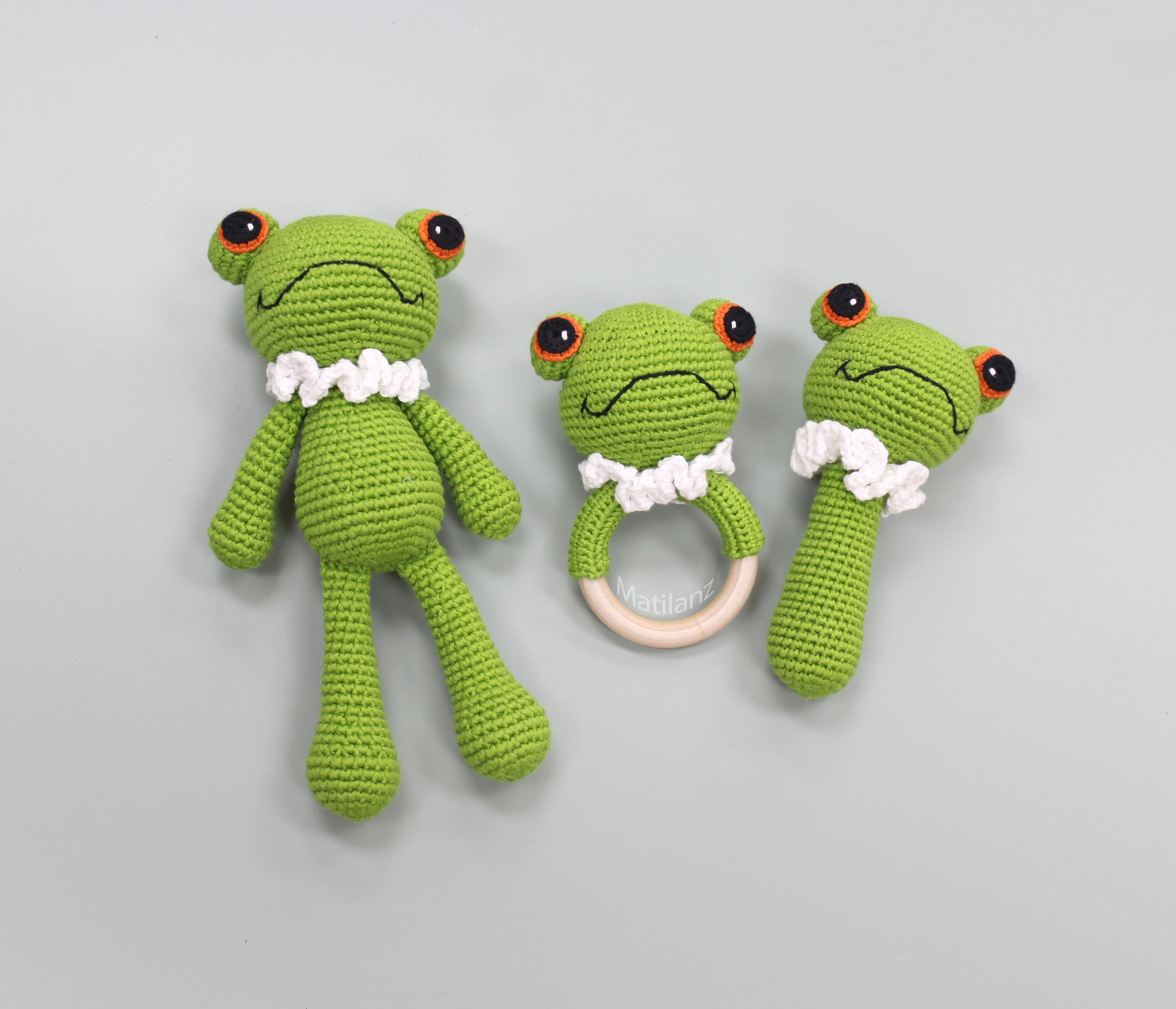 A handmade green crochet frog baby toy set with white collar, large eyes, and a wooden ring teether.
