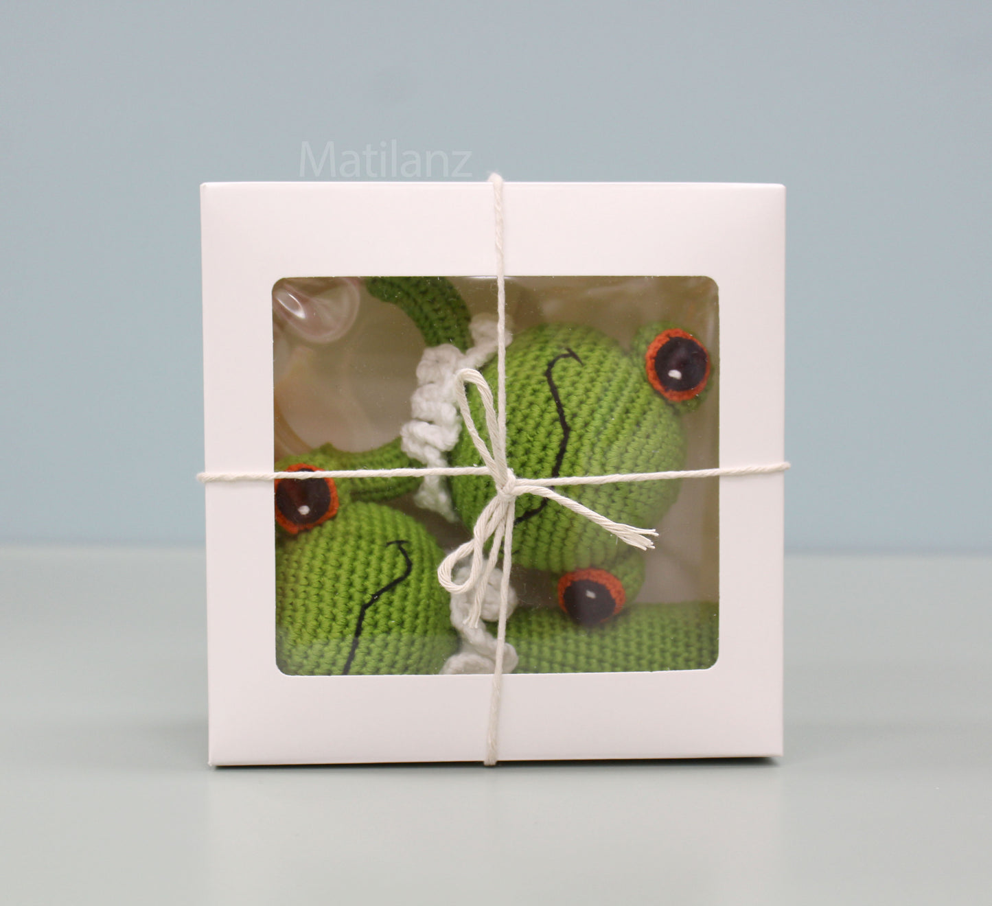 Handmade green crochet frog baby toy set with red eyes, nestled in a gift box tied with twine.