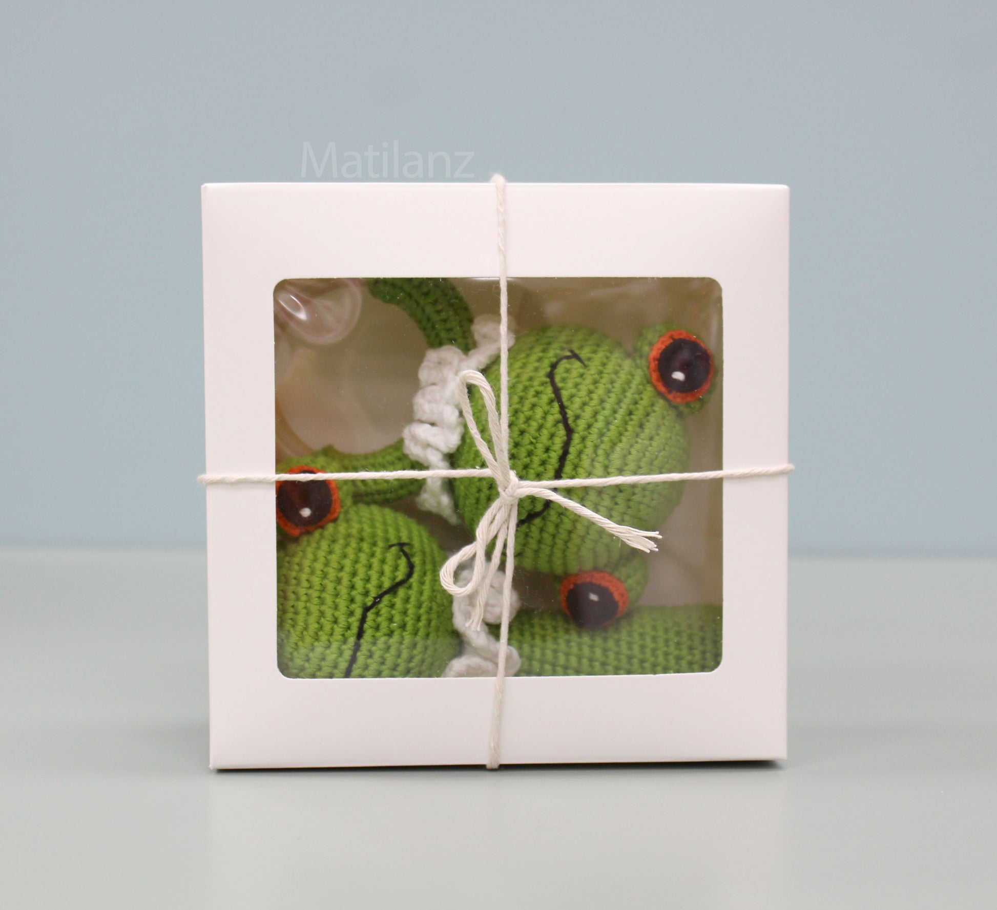 Handmade green crochet frog baby toy set with red eyes, nestled in a gift box tied with twine.