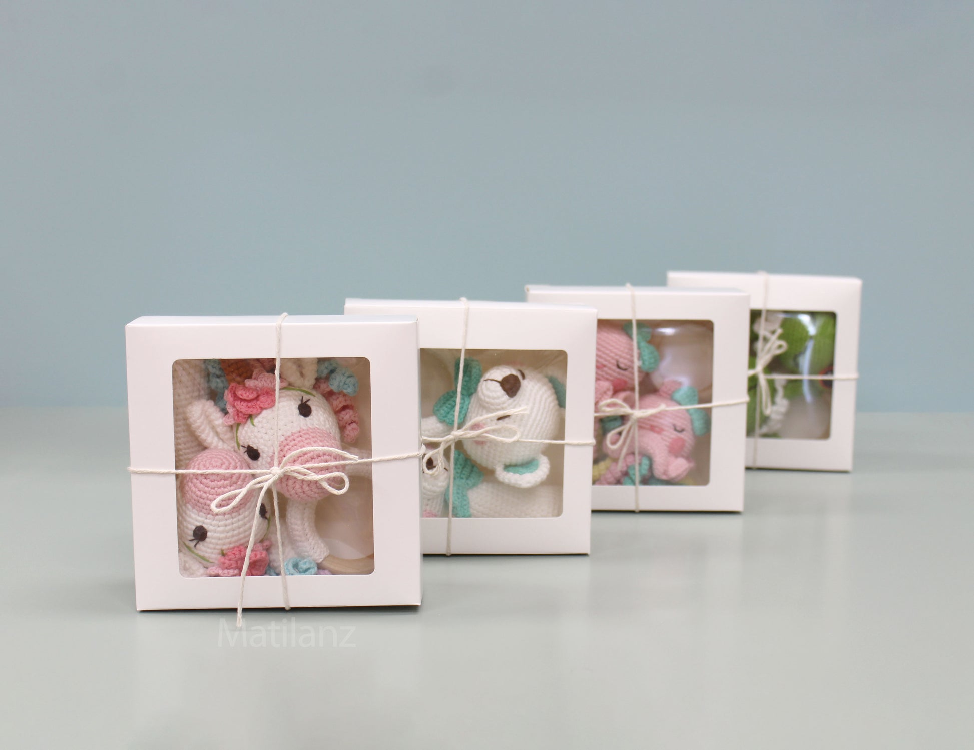 Handmade crochet bear rattles in soft pink and white, packed in clear gift boxes tied with twine.