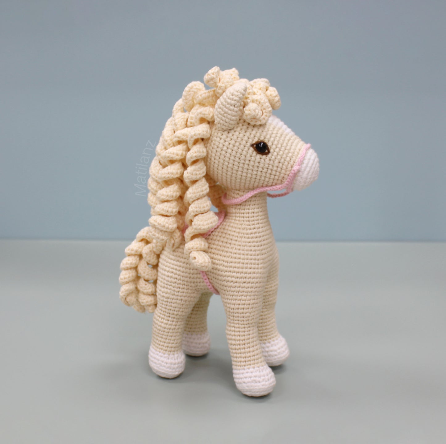 A handmade white crochet horse with a curly mane and tail, featuring pink accents and standing 10 inches tall.