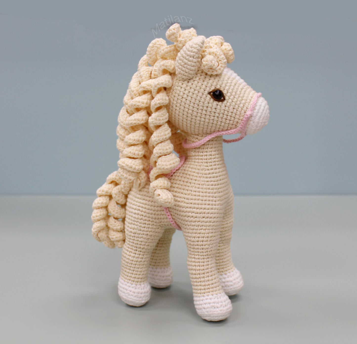 A handmade white cotton crochet horse with a curly mane and pink halter.