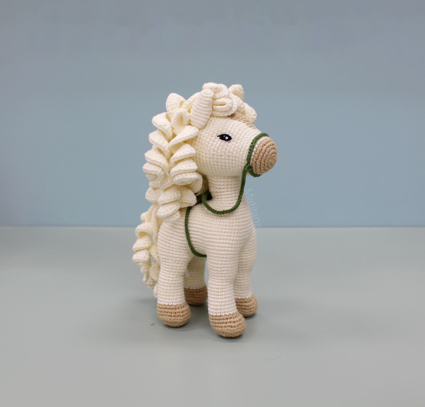 A white handmade crochet horse soft toy, 10 inches tall, with a fluffy mane and green reins.