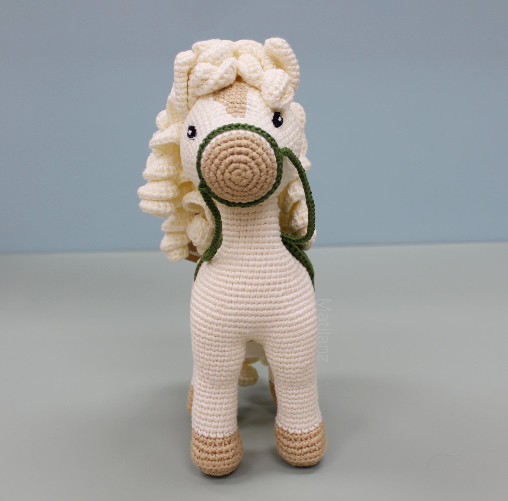 A handmade white cotton crochet horse toy, 10 inches tall, featuring a green reins and tan accents.
