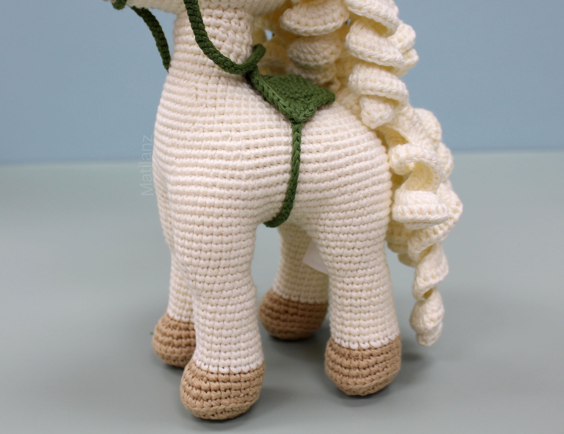 White handmade crochet horse soft toy with green saddle and brown hooves, 10 inches tall.
