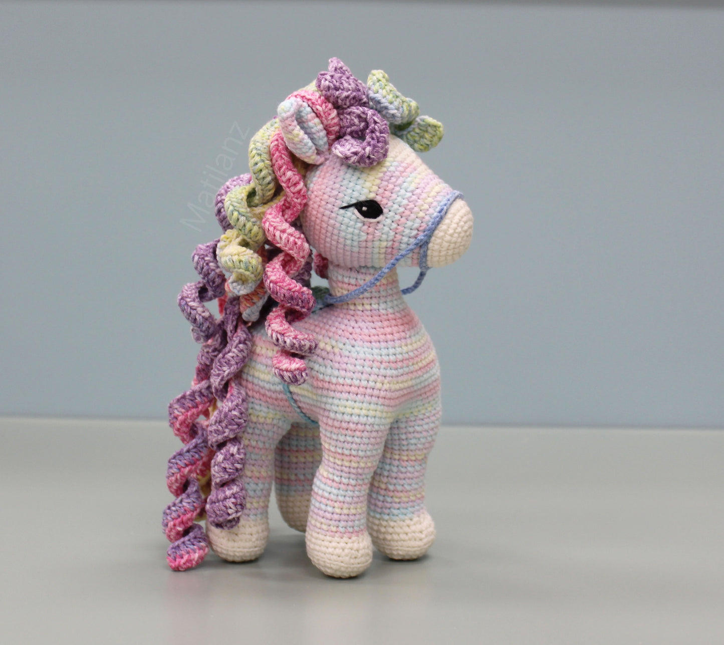 A handmade crocheted rainbow-colored horse with curly mane and tail, featuring detailed stitching and large expressive eyes.