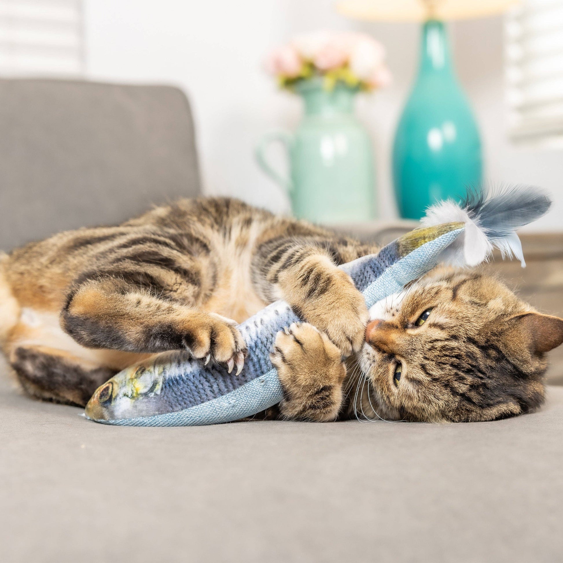 Plush blue fish cat toy with feathers and catnip, being playfully held by a tabby cat.