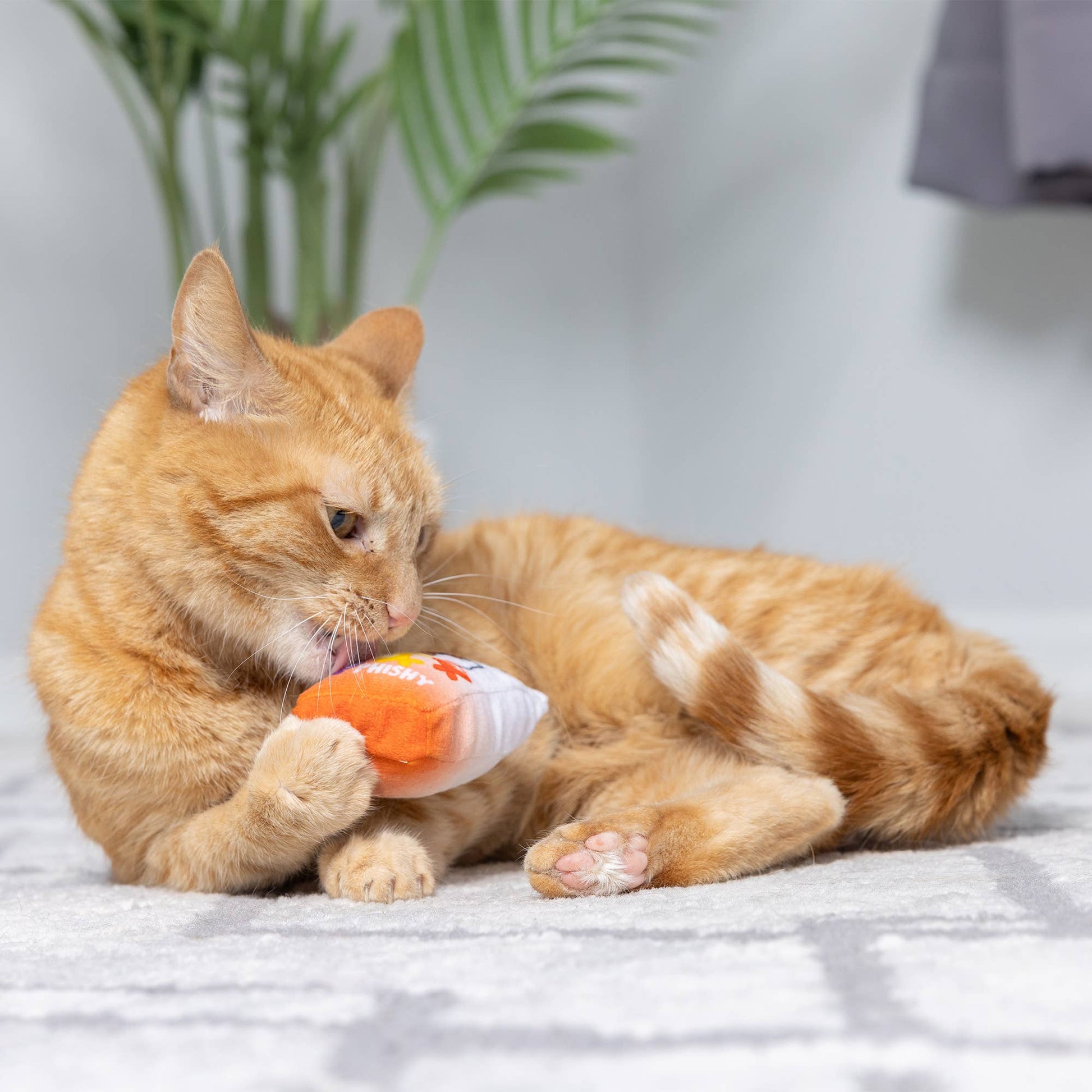 Orange plush cat toy shaped like a fish, being chewed by a cat.