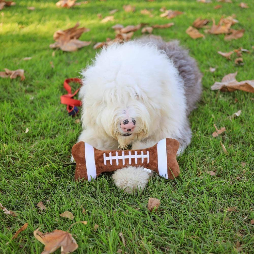 Brown and white football-shaped plush dog toy with durable stitching on grass.