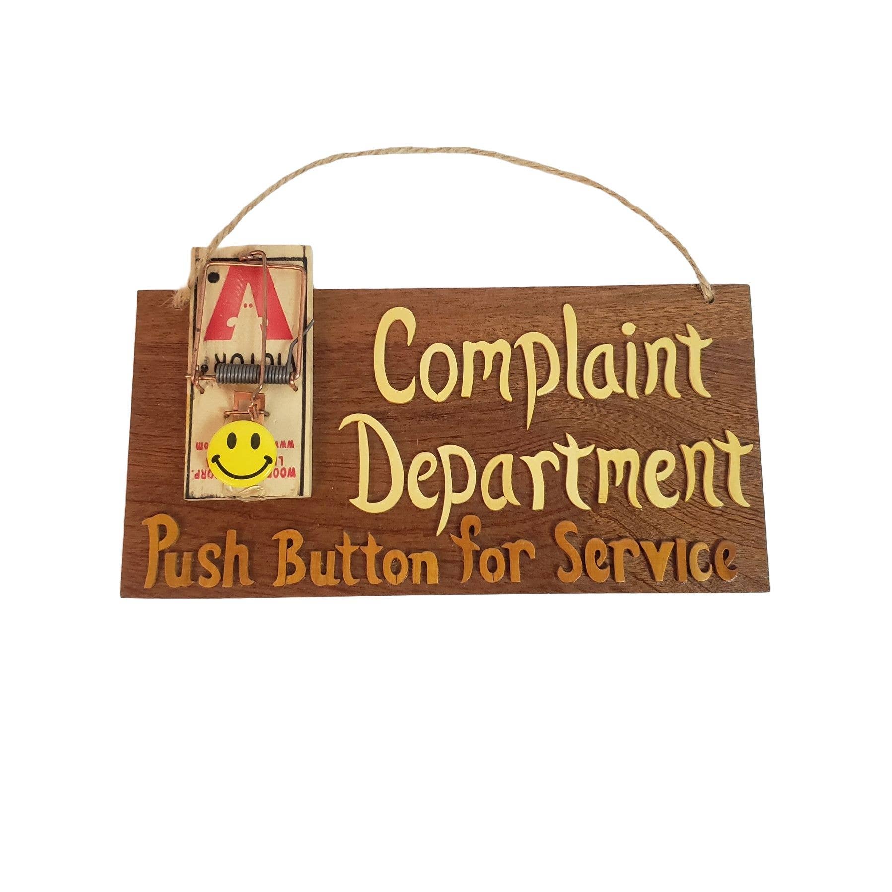 Wooden wall sign that reads “Complaint Department – Push Button for Service” with a mounted mouse trap featuring a smiley face.
