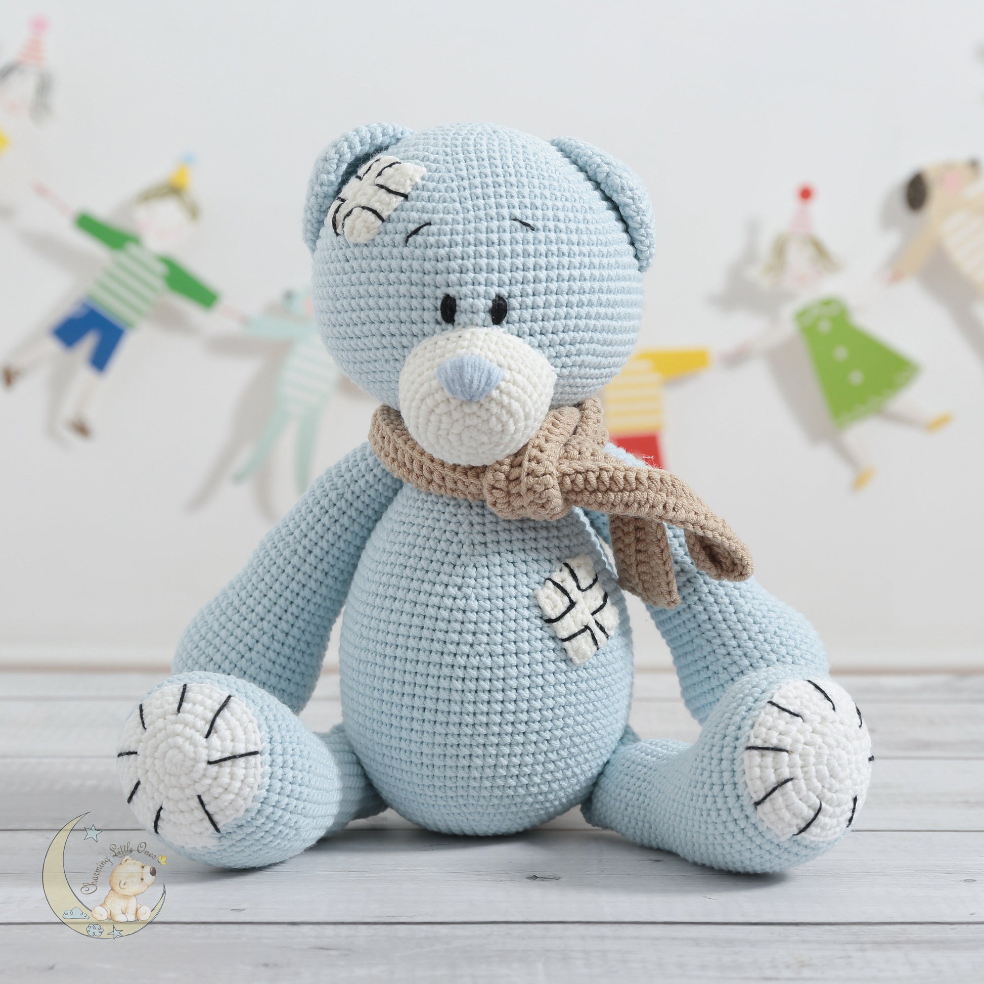 A light blue handmade crochet bear with a bandage on its head, wearing a beige scarf, and sitting on a wooden floor.
