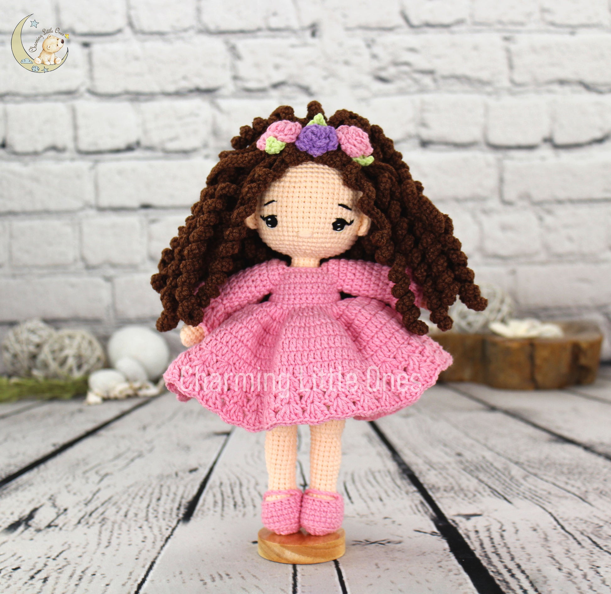 A handmade crochet doll with curly brown hair, pink dress, and flower crown, 10 inches tall.
