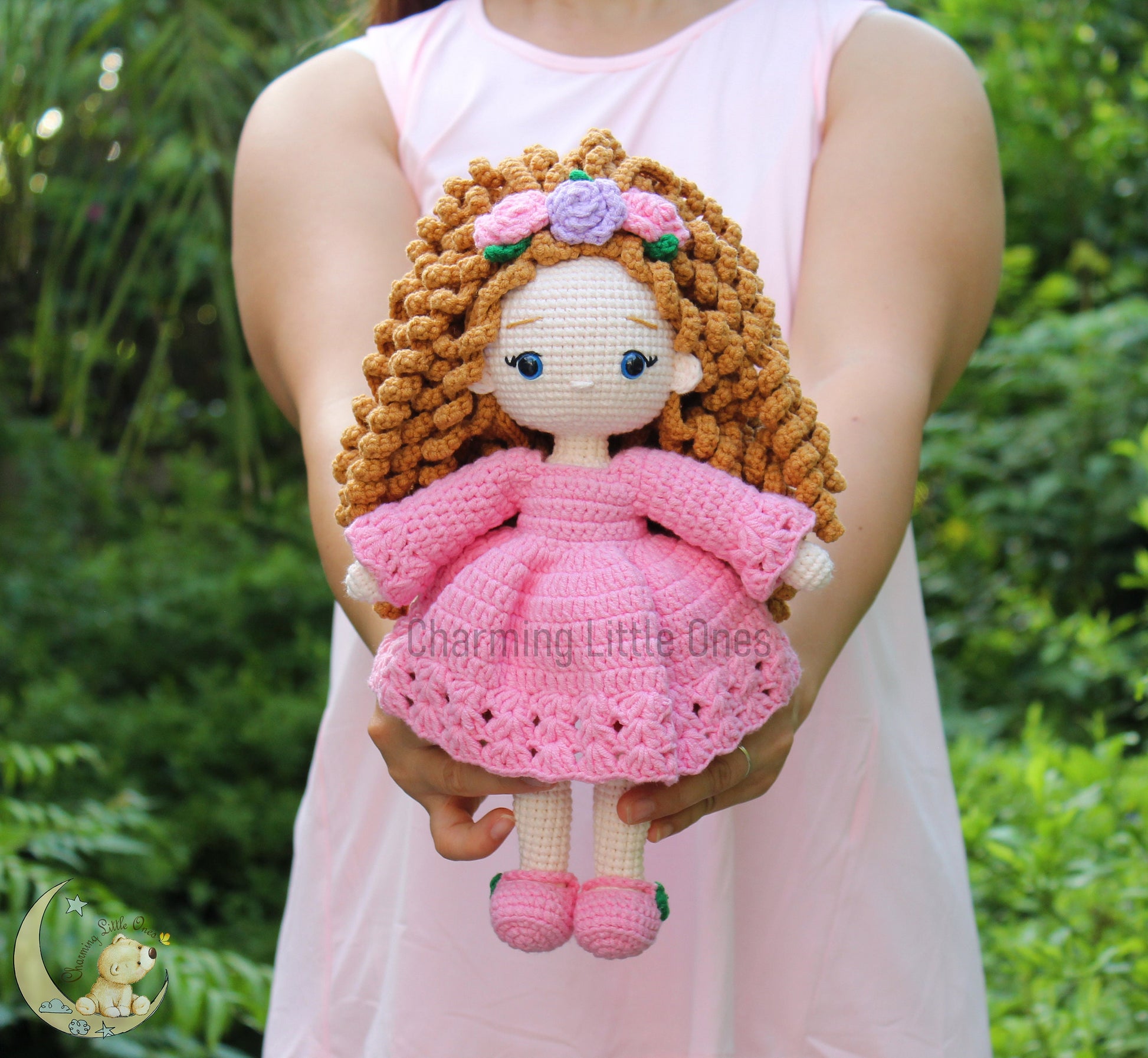 A handmade crochet doll with curly blonde hair, wearing a pink dress and floral headband.
