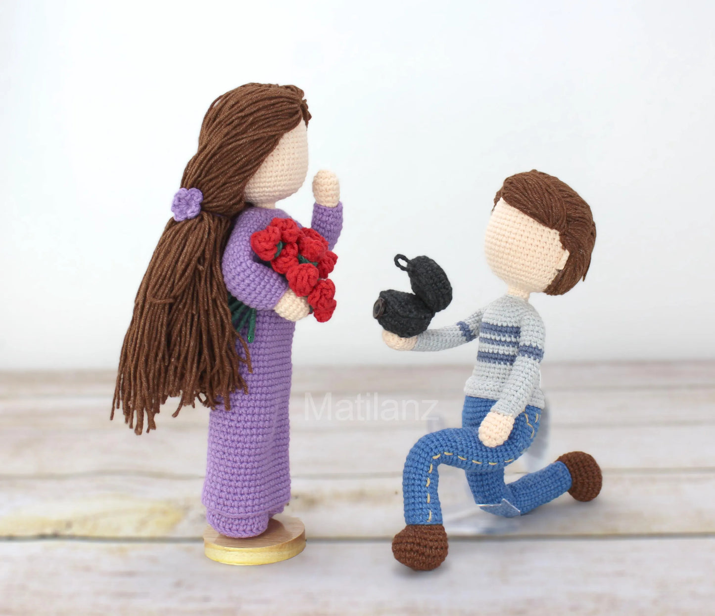 Crocheted dolls in a proposal scene, one holding flowers and the other kneeling with a ring box.