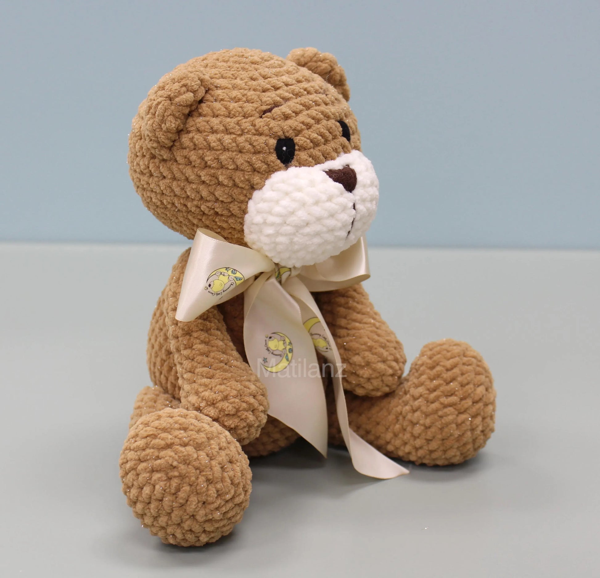 A beige crochet bear with embroidered eyes and a decorative bow featuring a moon and sun design.