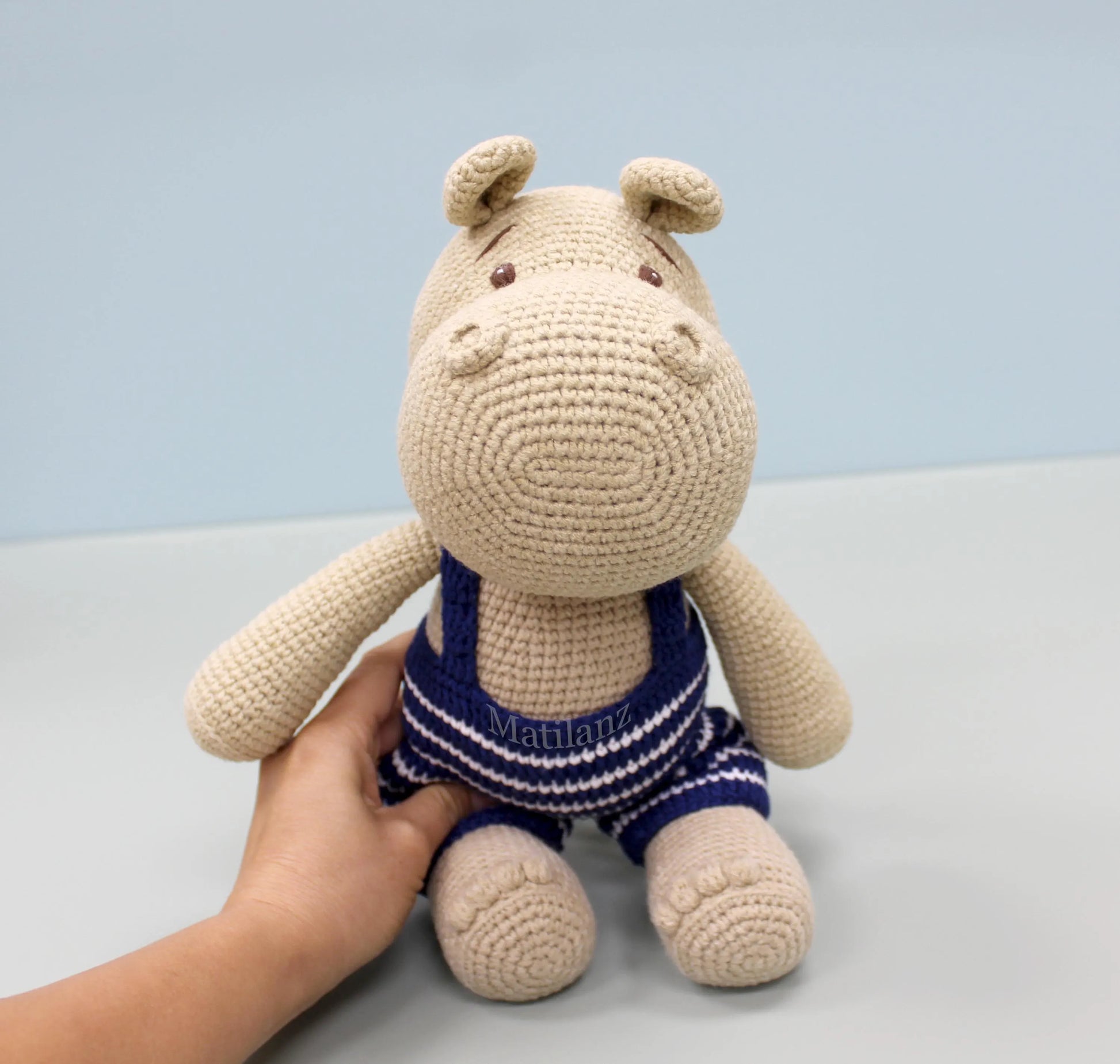 Crocheted beige hippo plushie wearing blue and white striped swim trunks.