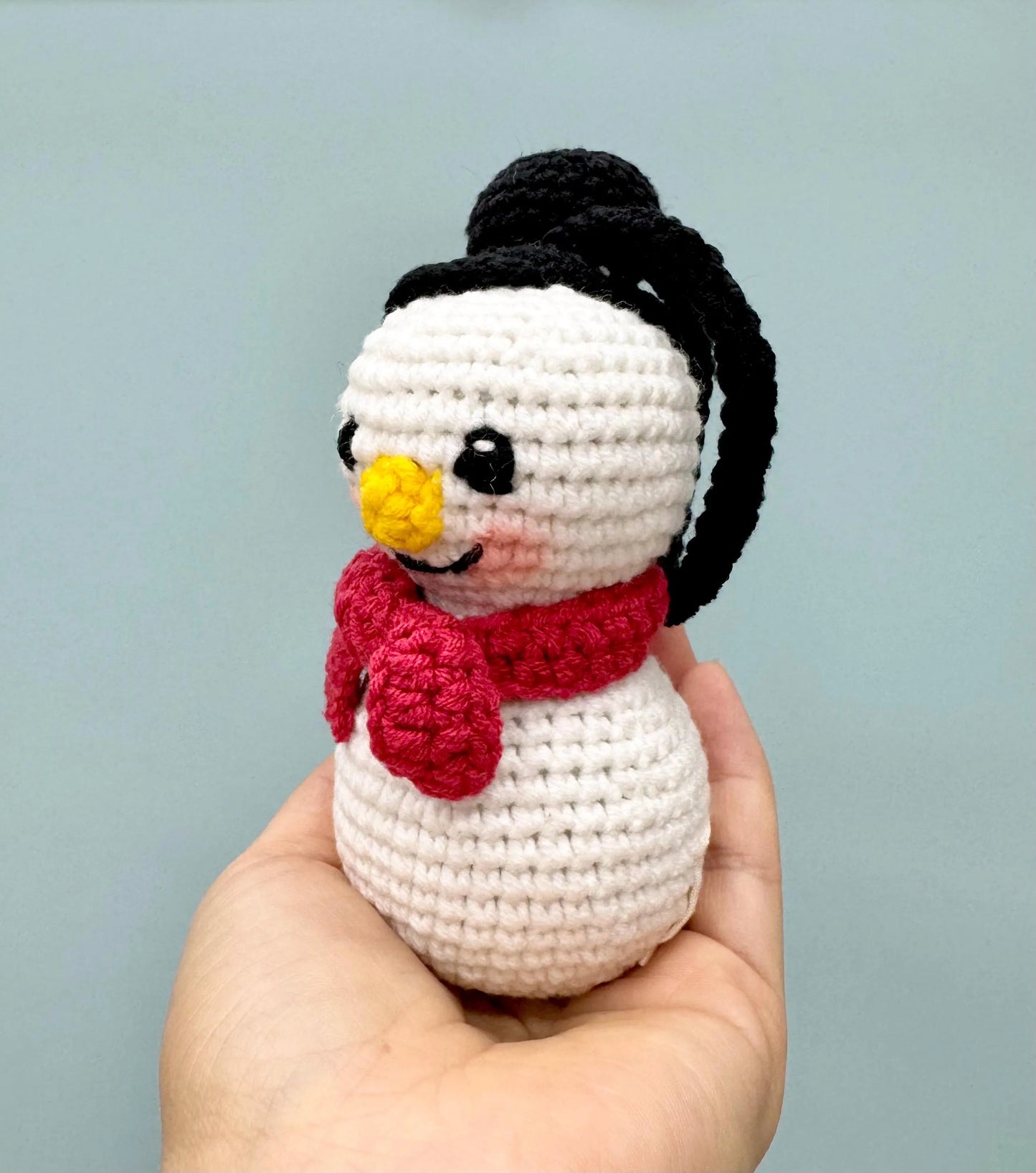 Crochet snowman ornament with a black hat and red scarf, featuring a yellow carrot nose and cheerful expression.