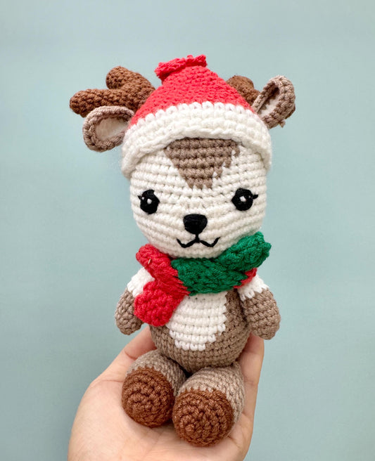 Crochet reindeer plush toy with brown and white fur, red Santa hat, and festive scarf.