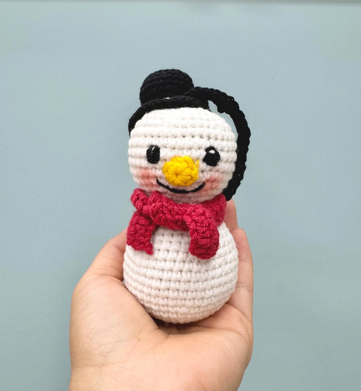 Crochet snowman ornament with a black hat and red scarf, featuring a cheerful yellow nose and rosy cheeks.