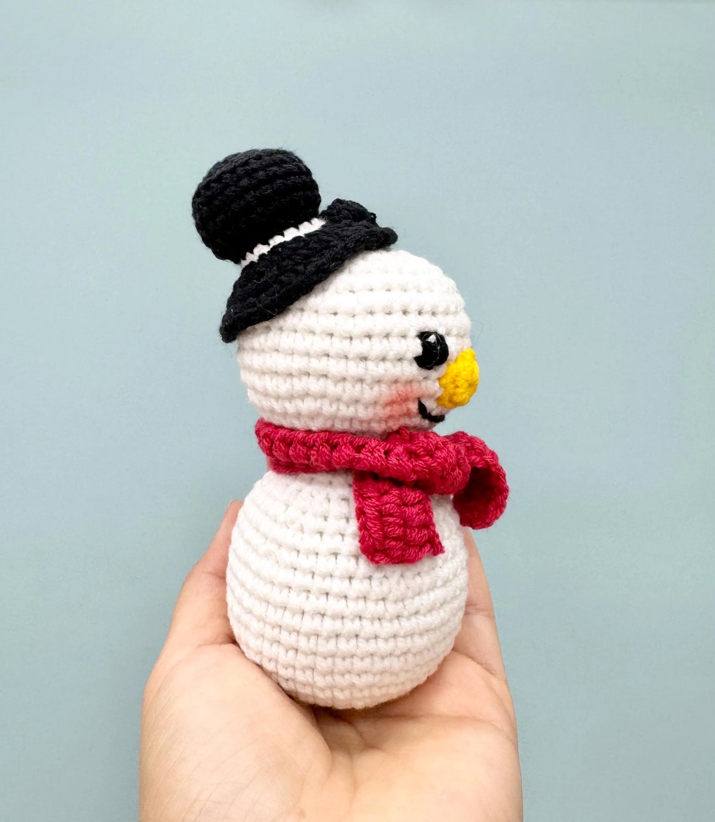 Crochet snowman ornament with a black top hat, red scarf, and cheerful yellow nose.
